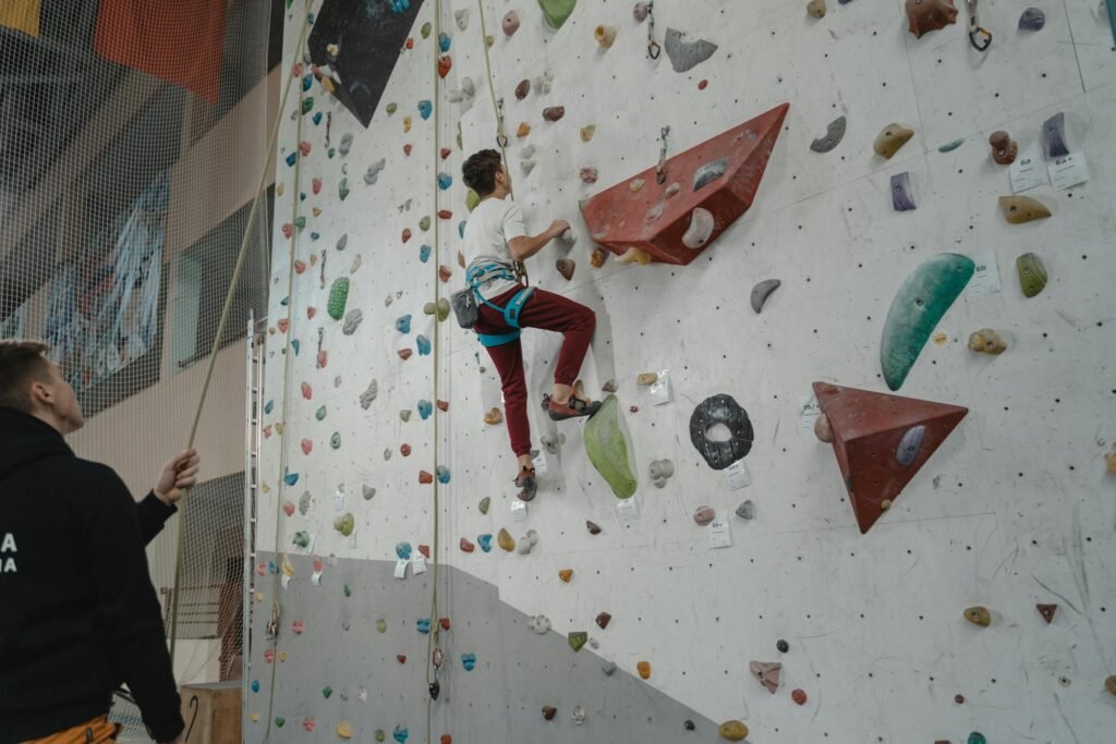 A young man practices climbing on an indoor rock wall, supervised by a trainer.