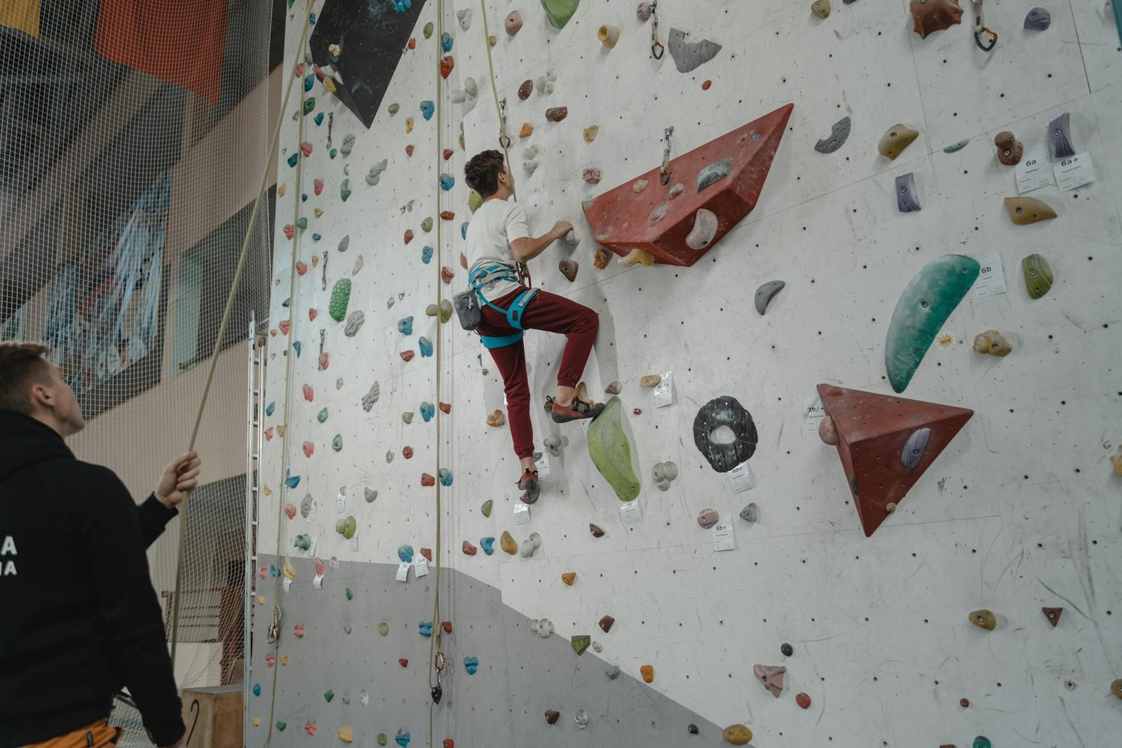 A young man practices climbing on an indoor rock wall, supervised by a trainer.