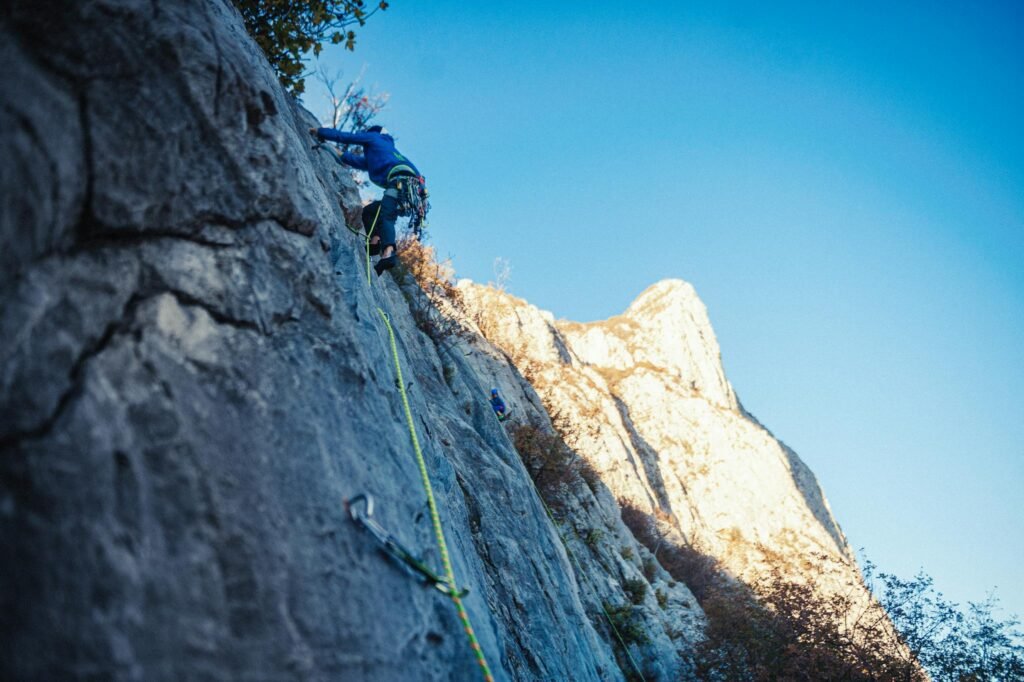 Adventurous rock climber ascending steep mountain face with safety gear in sunlight.