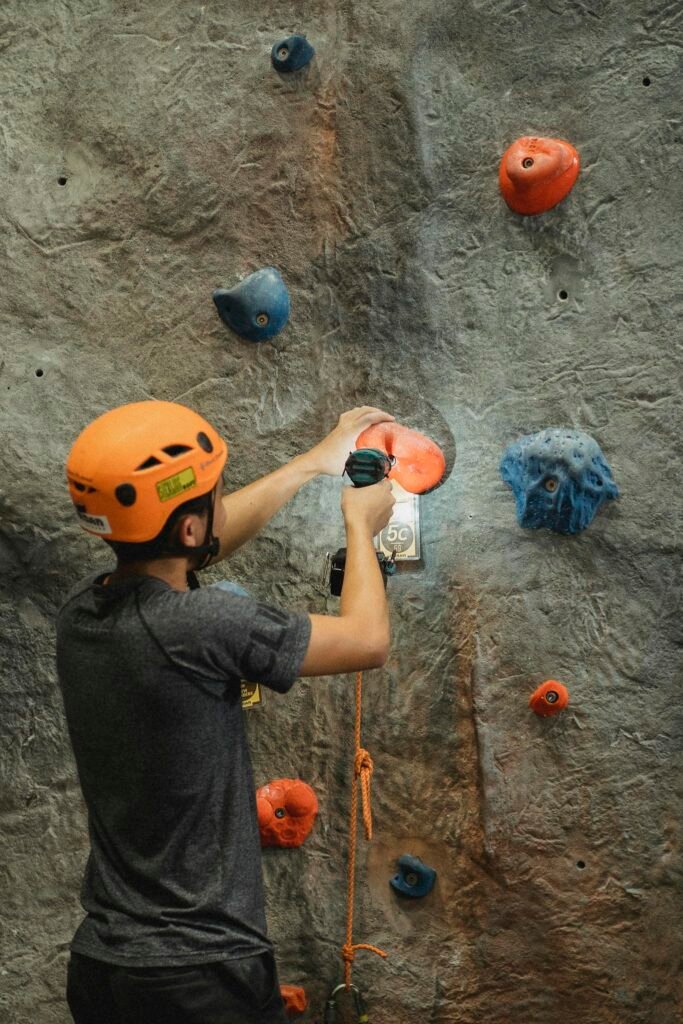 Back view of anonymous male mountaineer in protective helmet using electric screwdriver on grip of artificial rock